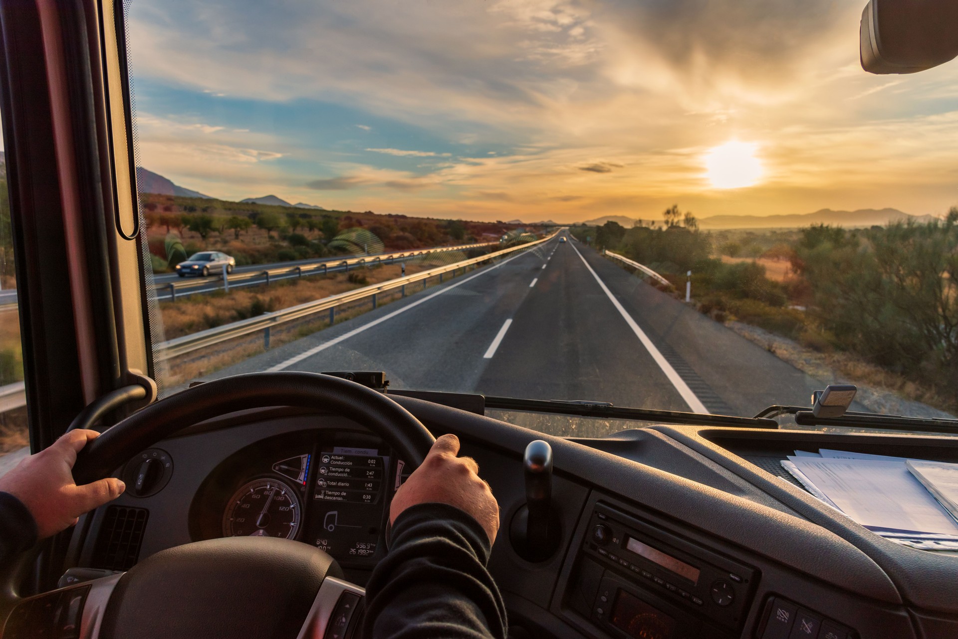 View from the driver's seat of a truck of the highway and a landscape of fields at dawn, with a dramatic sky.